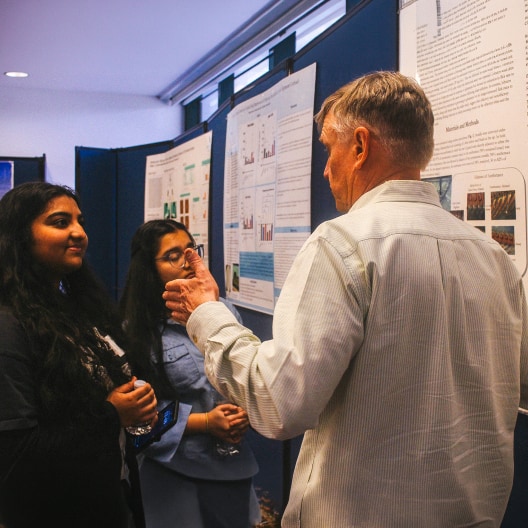 Two women and a man stand in front of scientific posters mounted on blue boards, engaging in a discussion. The man gestures with his hand while the women listen attentively.