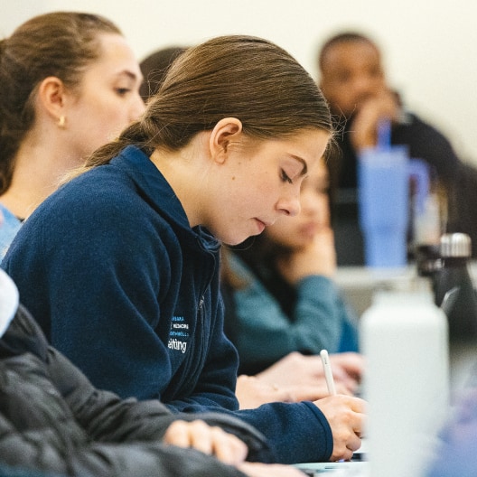 A young woman sits at a desk in a classroom, focused on writing in a notebook, surrounded by other students who are also engaged in their work.