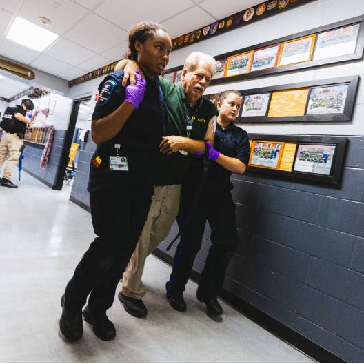 Two emergency responders assist an older man down a hallway, each supporting him by the arms. Both responders wear gloves and uniforms. Framed photos and awards decorate the hallway walls.