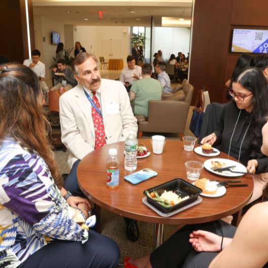 A group of people sit around a table having a meal and conversation in a well-lit, modern indoor setting with other groups in the background.