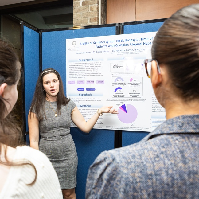 A woman stands in front of a scientific poster, explaining its content to two people. She gestures toward a section of the poster, which displays charts and text about lymph node biopsy research.