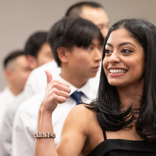 A woman with long black hair smiles broadly and gives a thumbs up in the foreground. Several people in white shirts stand in a line behind her, slightly out of focus.
