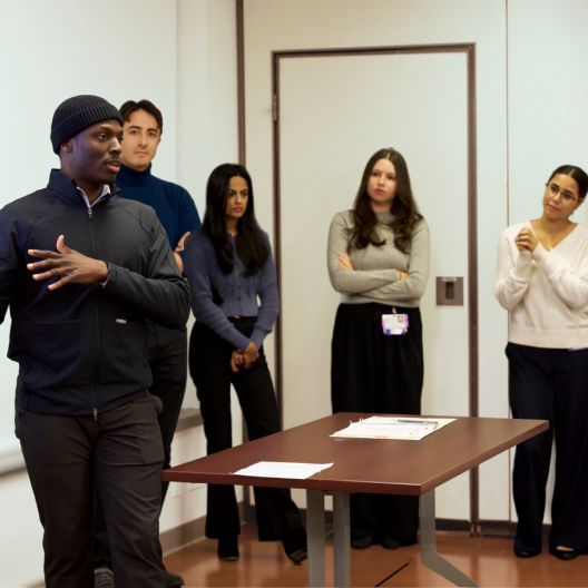 A man in a black jacket and beanie speaks to four people standing by a table in a room, with papers placed on the table. The group listens attentively.