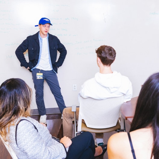 A man in a blue cap stands and speaks to three seated people in front of a whiteboard with writing on it, in a casual classroom or meeting setting.