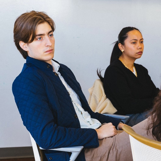 Two people sit attentively in chairs during what appears to be a meeting or class. The man in front wears a blue jacket, and the woman behind him wears a black sweater and gold necklace, both looking focused.
