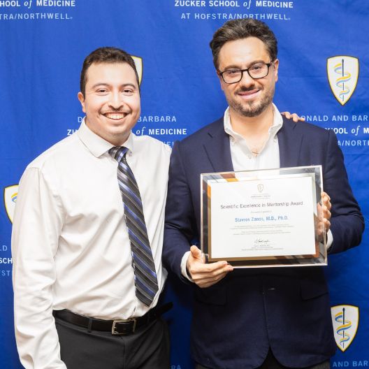 Two men in dress shirts stand smiling in front of a blue backdrop. One man holds a framed Scientific Excellence in Mentorship Award. The backdrop reads Zucker School of Medicine at Hofstra/Northwell..