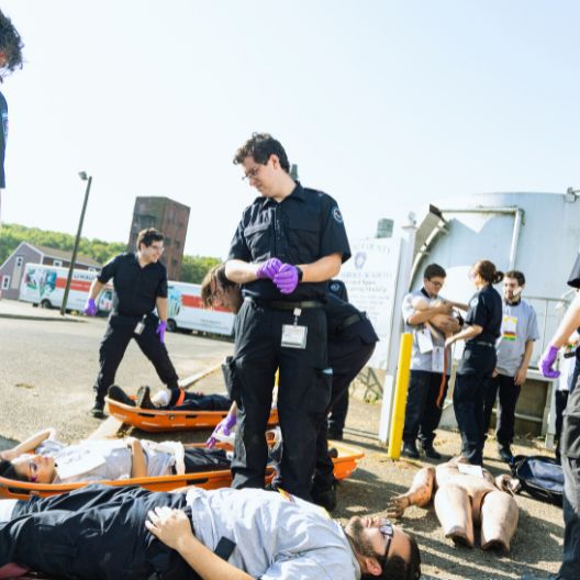Emergency responders wearing black uniforms and purple gloves attend to people lying on stretchers outdoors during a daylight drill or training exercise. Signs and buildings are visible in the background.