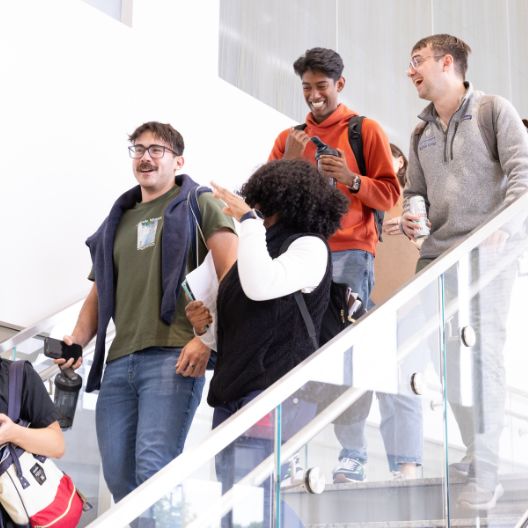 Four young adults walk down a glass staircase, laughing and talking together. One person holds a camera, and another carries a can. Everyone appears to be enjoying themselves in a bright indoor setting.