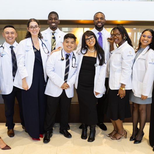 Eight smiling medical professionals in white coats and business attire stand together indoors, posing for a group photo. Some wear stethoscopes around their necks. They appear happy and confident.