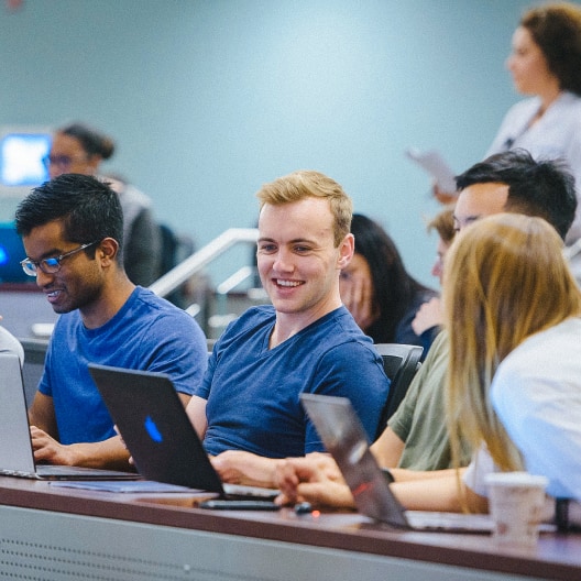Four students sit together in a classroom, using laptops and talking. One student in the center is smiling, while others are engaged in conversation. There are more people and a woman standing in the blurred background.