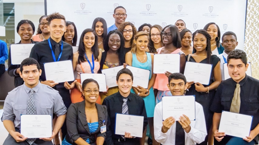A group of young adults pose together indoors, smiling and holding certificates. They are dressed in business or semi-formal attire, standing in front of a branded backdrop at an award or graduation event.