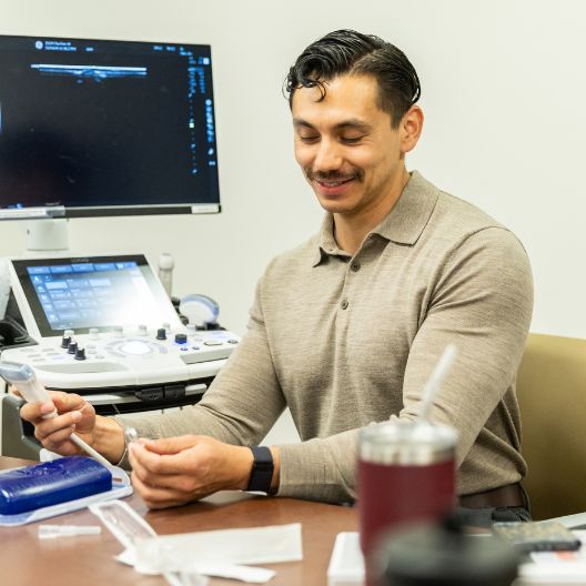Medical student in a beige shirt sits at a desk with medical equipment, holding an ultrasound probe. A monitor and various medical tools are visible around him.