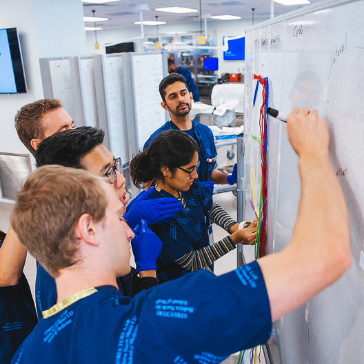A group of people in blue scrubs gather around a whiteboard, collaborating and taking notes in a bright, modern room with workstations in the background.