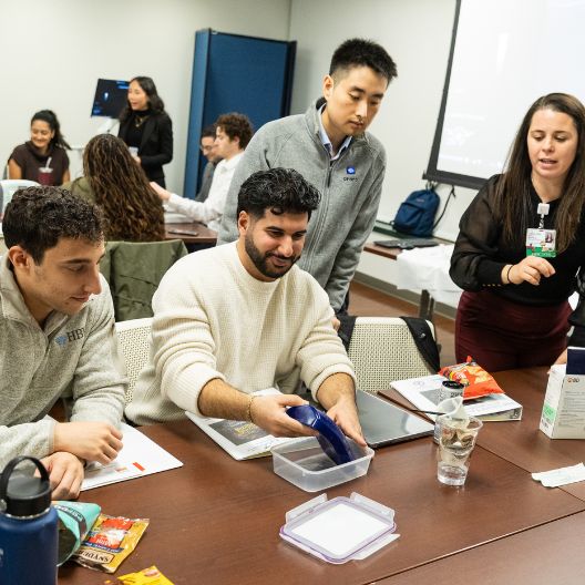 Medical students gather around a table with snacks, notebooks, and boxes. One man pours liquid from a blue bottle into a container while others watch and discuss. A presentation is visible in the background.