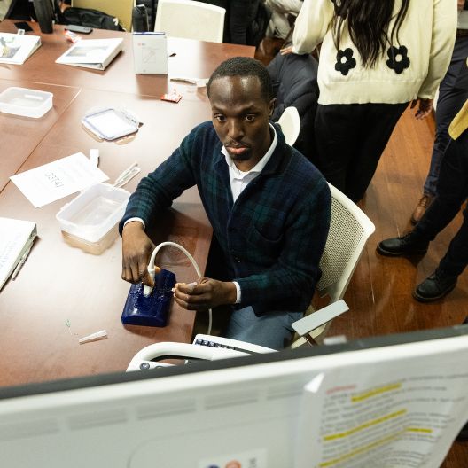 Med student sits at a table using a medical simulation device, with papers, containers, and syringes nearby. Other people stand around in the background on a wooden floor.