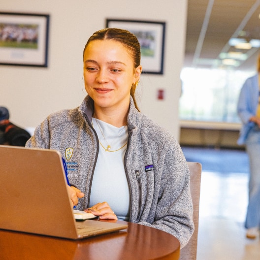 A woman sits at a table, smiling while working on a laptop and taking notes with a pen. She wears a light blue shirt and gray fleece jacket in a bright, indoor setting with blurred people and framed photos in the background.