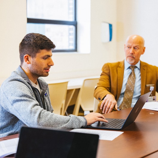 Two men sit at a table in an office setting. One is typing on a laptop while the other, dressed in a suit, looks on attentively. Papers and another laptop are visible on the table.