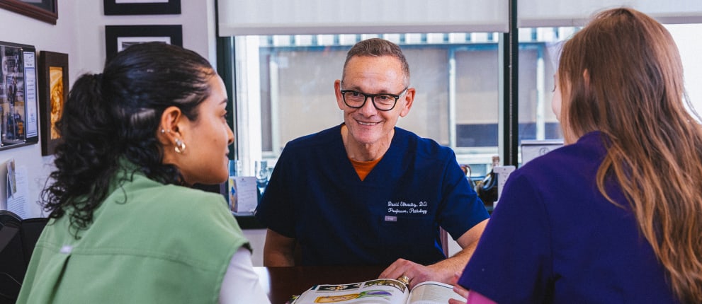 A doctor in blue scrubs smiles while talking to two women at his office desk. An open book lies on the table, and framed photos hang on the wall in the background.