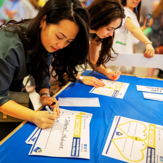 Two women lean over a blue table, writing on large cards with heart designs. Other people and papers are visible in the background. The setting appears to be a community or networking event.
