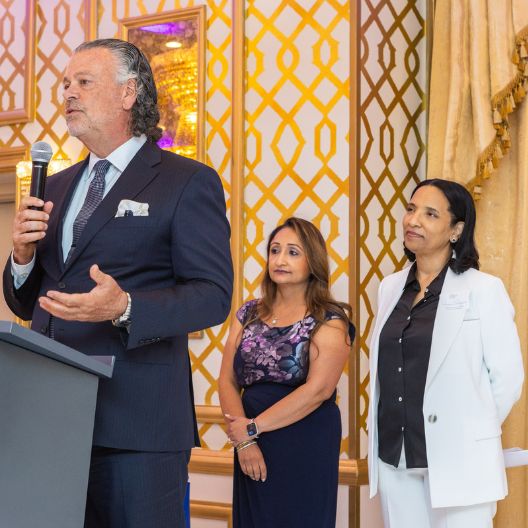 A man in a suit speaks at a podium with a microphone, while two women stand beside him, listening attentively. The background features gold-patterned walls and elegant curtains.