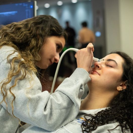 A woman uses a handheld medical device to scan another womans face as she reclines with her eyes closed, likely during a medical or cosmetic procedure. Both are wearing casual gray clothing.