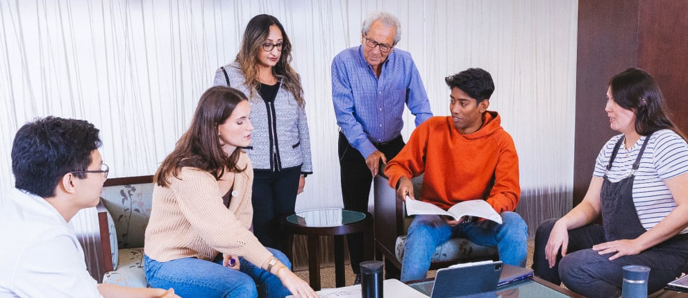 A group of six people sit and stand around a table, engaged in discussion. One person holds an open book, while a laptop and papers are on the table. They appear to be collaborating or studying together.