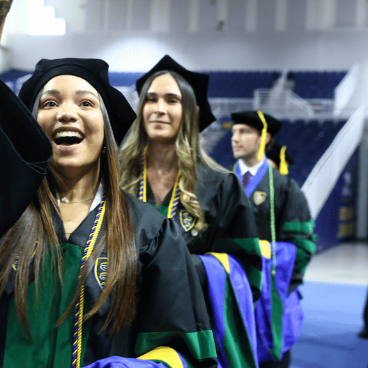 A group of graduates in caps and gowns smile and celebrate while standing in line at an indoor graduation ceremony.