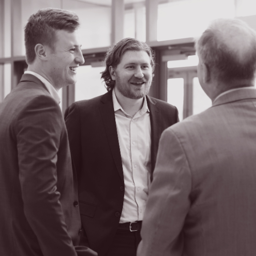 Three men in suits stand together indoors, engaged in conversation and smiling. Two face the camera, while the third is seen from behind. The atmosphere appears professional and friendly.