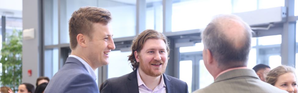 Three men in business attire talk and smile together indoors, with large windows in the background. Other people are visible in the blurred background of the bright, modern space.