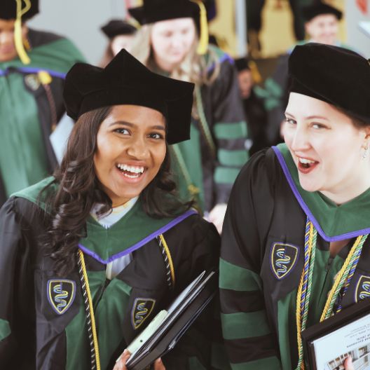 Two graduates in black and green academic robes smile and hold diplomas at a graduation ceremony, surrounded by others in similar attire.
