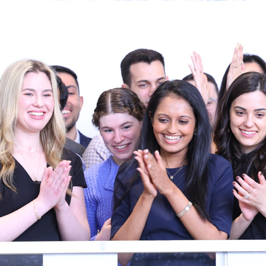 A group of people stand close together, smiling and clapping. Several women in the front are especially joyful, applauding enthusiastically. The background is bright and white.