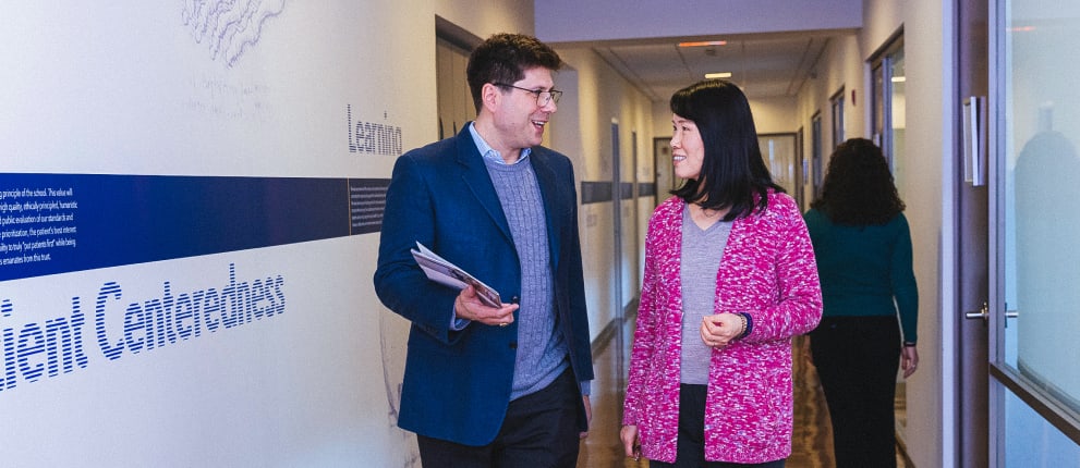 A man in a blue blazer and glasses walks with a woman in a pink cardigan down a hallway, smiling and holding papers, while another person walks ahead of them.