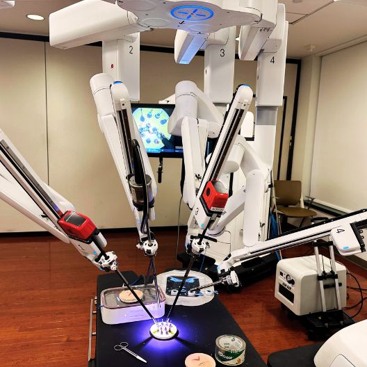 A robotic surgical system with multiple articulated arms operates on a simulated patient, with a monitor displaying images and medical tools nearby in a clinical room.