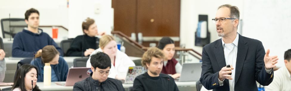 A man in a suit speaks at the front of a classroom while students listen and take notes on laptops.