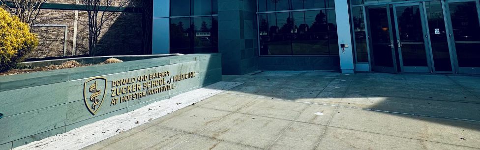 Entrance to a building with glass doors. A sign on the left reads, Donald and Barbara Zucker School of Medicine at Hofstra/Northwell. Shrubs and a brick wall are in the background.