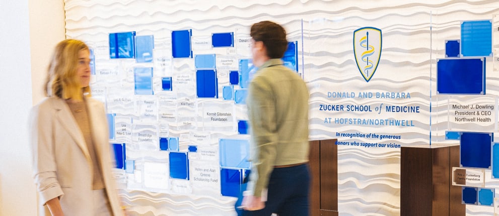 Two people walk past a donor recognition wall at the Zucker School of Medicine at Hofstra/Northwell, featuring blue and clear plaques and the school’s name and emblem.