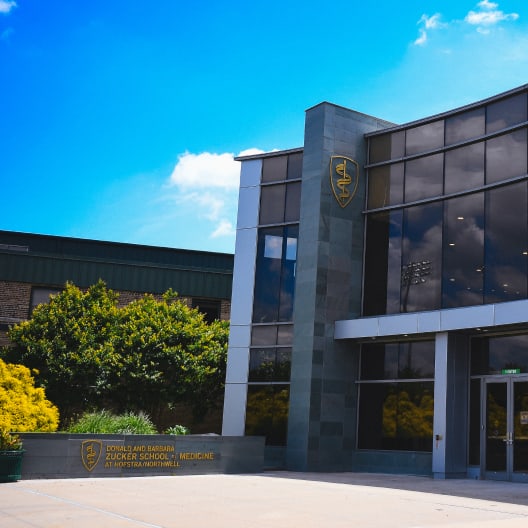 Modern glass building with reflective windows and a crest, labeled Zucker School of Medicine at Hofstra/Northwell, surrounded by green bushes under a bright blue sky with scattered clouds.