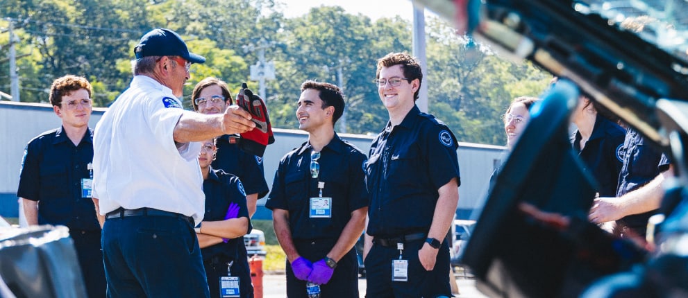 A group of people in dark uniforms listen to an instructor, who is gesturing with a red object, during an outdoor training session beside a vehicle on a sunny day.