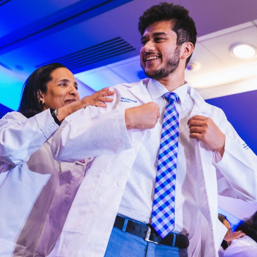 A smiling young man in a dress shirt and checkered tie is helped into a white coat by a woman during a formal ceremony. Both appear happy, and the setting has bright indoor lighting with a blue accent.