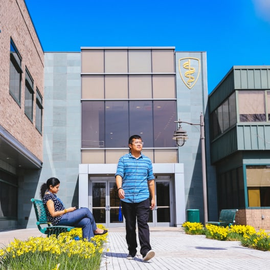 A man walks on a pathway lined with yellow flowers outside a modern building with large windows, while a woman sits on a bench looking at her phone. A medical caduceus symbol is displayed on the building.