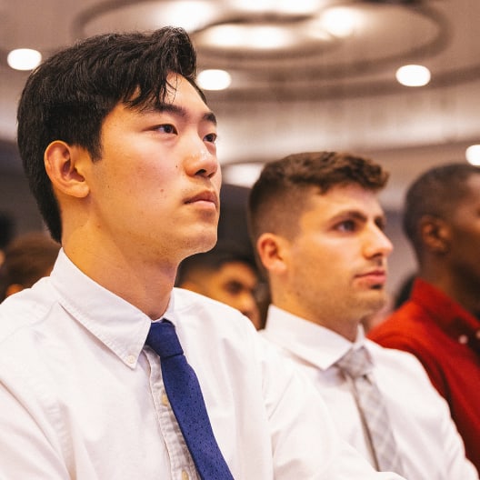 Three men in dress shirts and ties sit attentively in an audience, focused on something ahead of them. The background is softly lit with circular ceiling lights visible.