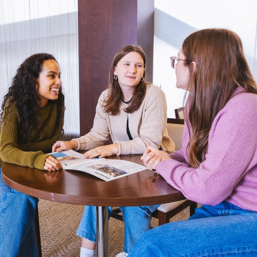 Three women sit around a round wooden table, smiling and talking. One gestures to an open magazine on the table. They appear to be having a friendly and engaged conversation in a well-lit room.
