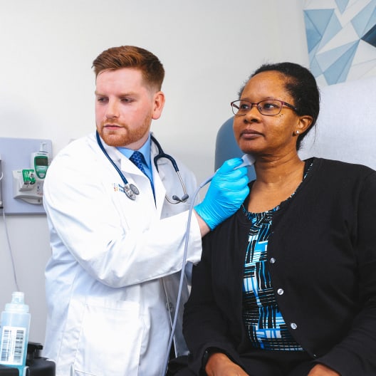 A doctor wearing a white coat and blue gloves uses a medical device to examine a seated woman’s neck in a clinical setting. The woman wears glasses and sits calmly during the examination.