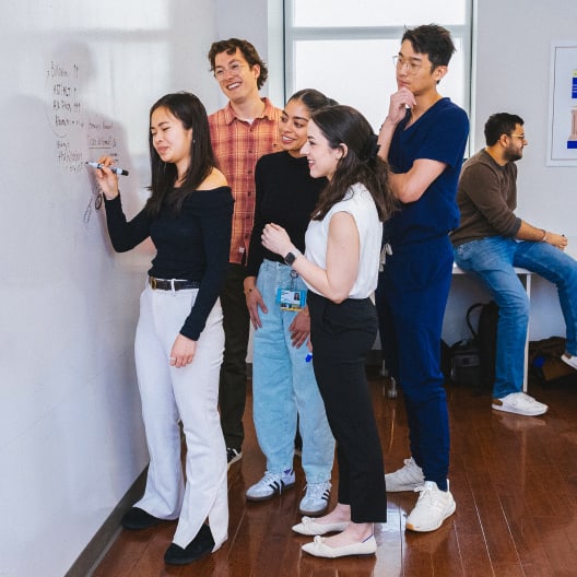 Five young adults stand by a whiteboard, with one woman writing while the others watch and smile. Another person sits in the background at a table, looking at a laptop. The setting appears to be a casual classroom or office.