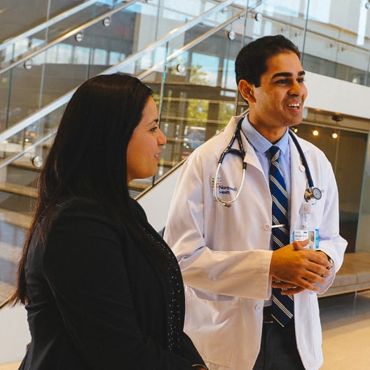 A doctor in a white coat smiles and talks with two women in business attire inside a modern building with large windows and a glass staircase. One woman holds a folder.