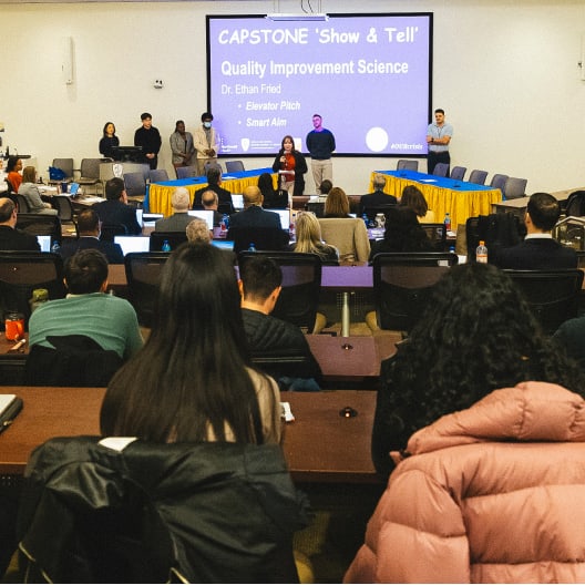 A presenter speaks at the front of a conference room during a CAPSTONE Show & Tell event on Quality Improvement Science. Audience members are seated, with a presentation projected on a screen behind the speakers.