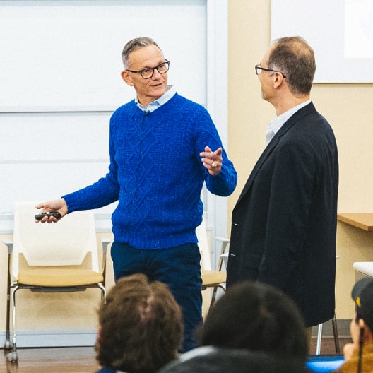 Two men stand at the front of a classroom, engaged in conversation. One gestures while speaking, holding a remote, as several people seated in the audience listen attentively.