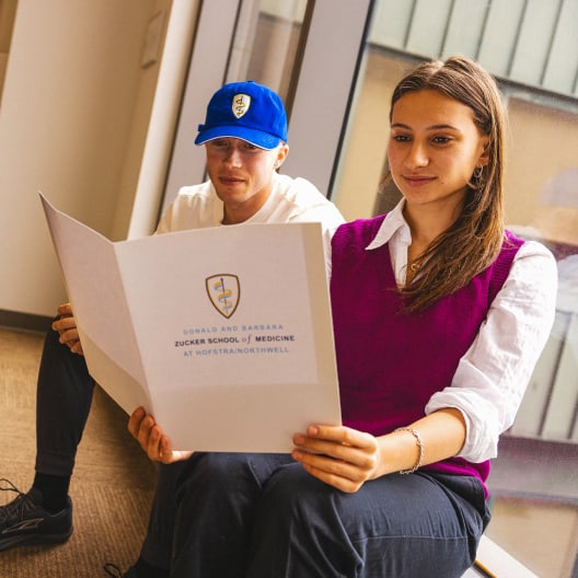 Two people sit by a window reading a folder labeled “Zucker School of Medicine at Hofstra/Northwell.” One wears a blue hat and white shirt; the other wears a white shirt with a purple vest and smiles at the folder.