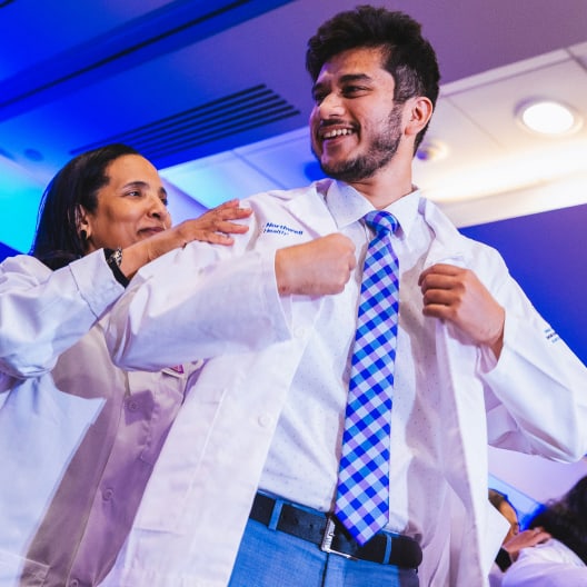 A smiling young man in a blue checkered tie is helped by a woman as he puts on a white coat, likely at a medical or graduation ceremony, under bright indoor lighting.