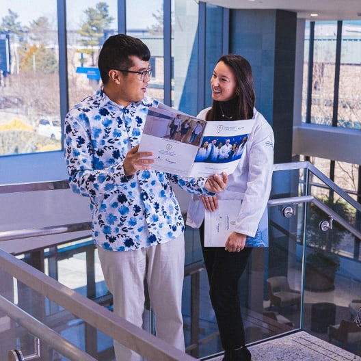 Two people stand on a staircase indoors, looking at a brochure together. One person wears a floral shirt and glasses, and the other wears a white coat and smiles. Large windows reveal trees and sunlight outside.